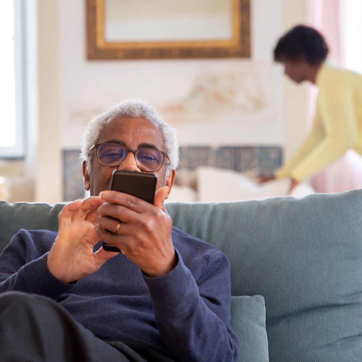Older man on the sofa while a care worker makes the bed at home